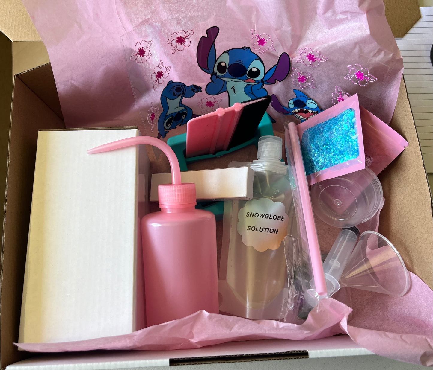 A box containing a snow globe starter kit with various tools and materials including a pink funnel, a syringe solution, a weed tool, a UV torch wrap, straws, and a resin mixing tub.